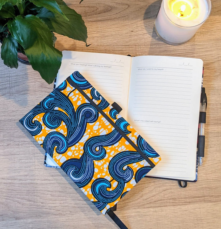 yellow and blue wellness journal on top of open wellness journal with self care prompts, on a wooden table next to a lit candle and green plant